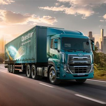 Blou vragmotor ry op oop pad met stad se skyline en bome in die agtergrond.