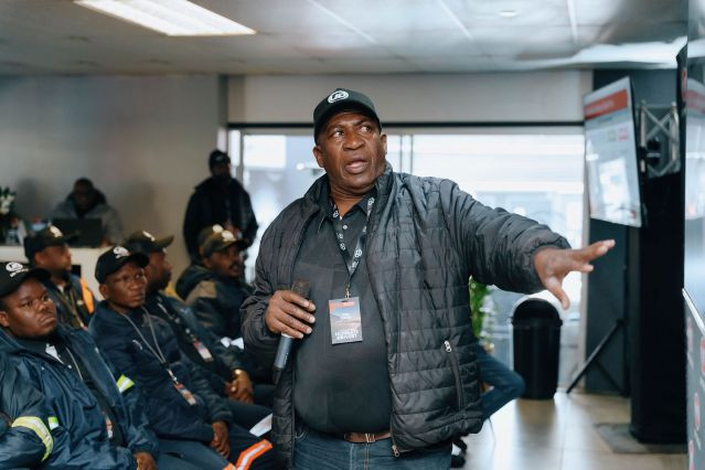 Man in a black jacket giving a presentation to seated people in a room.
