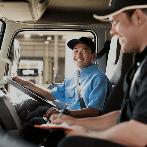 Two men in a truck cabin, one driving and smiling, the other writing on a clipboard.