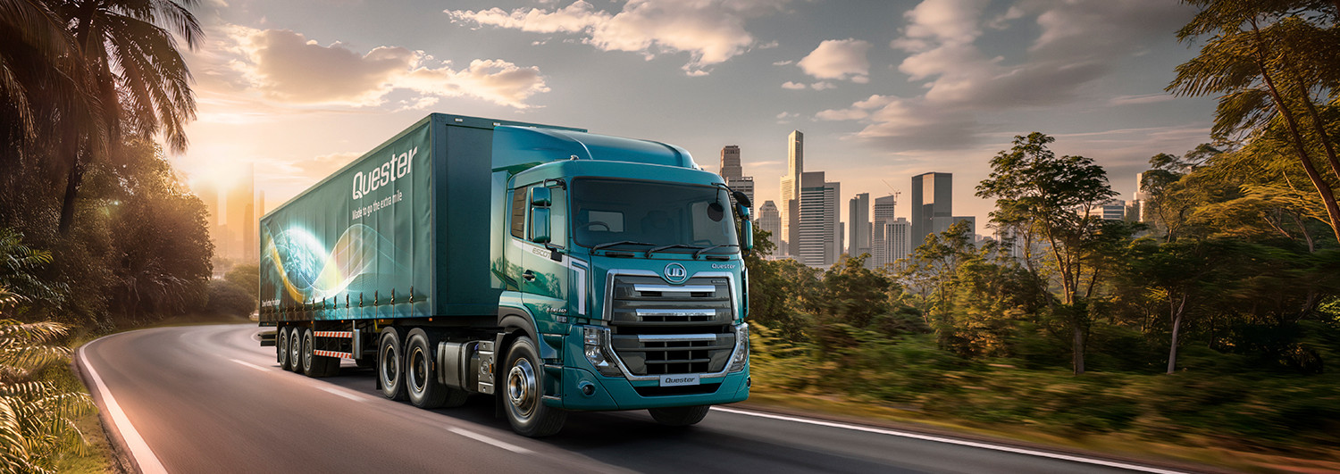 Blue semi-truck driving on a highway with a city skyline in the background at sunset.