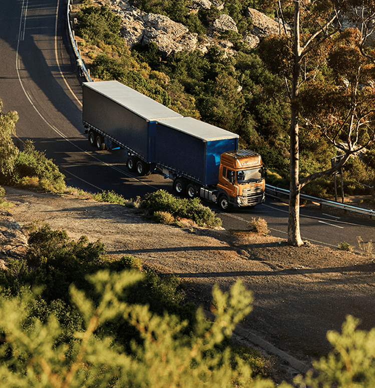 Lori treler biru memandu di jalan berliku di kawasan berbukit dengan pokok-pokok.