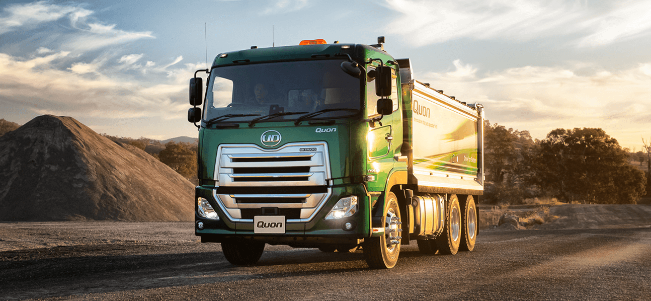 Green dump truck on a gravel road at sunset, sand piles in the background.